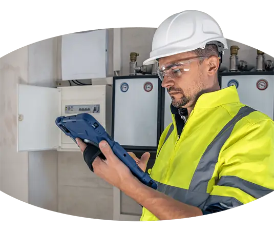 Ein Mann mit Schutzhelm benutzt ein Tablet, während er eine Schalttafel auf einer Kranführer-Baustelle inspiziert.