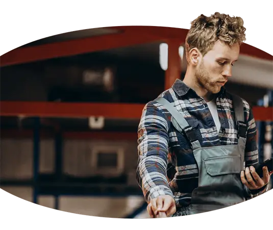 Ein Mann im Overall, der in einem Lagerhaus ein Mobiltelefon benutzt, während er Kranführerjobs überwacht.